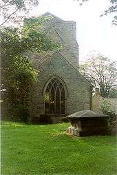 Photograph showing an old stone church with a short wide tower. The view is taken from a graveyard, there is a large tomb stone in the foreground and the church is surrounded by trees.
