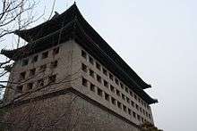 A tall rectangular stone building with a Chinese-style roof seen from near its base with some bare tree branches in front