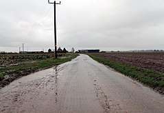 A completely flat landscape.  A wet, muddy road stretches in the distance, dead straight, flanked by empty, wet fields.  A wooden electricity pole is a sudden vertical element.  The wires follow the road down to some farm buildings in the middle distance