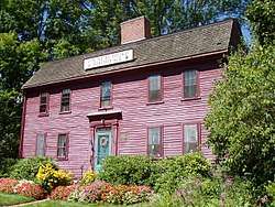 A red two story colonial house with wooden roof. Flowers have been planted on either side of the front door.