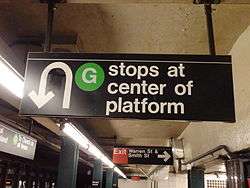 An overhead sign at an underground "G" train station. The sign's text says, "'G' stops at center of platform". There is a U-turn arrow icon on the left side of the sign, next to the text.