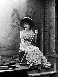Full length monochrome photograph of a woman seated on a low stool in front of a painting and facing slightly to her right. She wears a high-waisted flowered dress with a solid colour sash and a broad-brimmed hat decorated with flowers and holds a long walking stick.