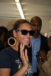 A woman wearing a printed T-shirt, sunglasses and hoop earrings. She is smiling and waving, and various people are in the background.