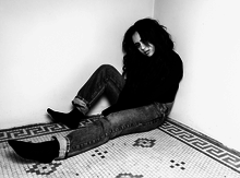  Picture of a young, brunette woman who is sat on the floor and rests on a concrete wall. She wears jeans and a dark shirt.