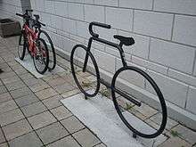 Two bicycle racks shaped like stylized bicycles, in metal with a gloss black finish, each set in a rectangle of concrete within a floor of small ceramic tiles, in front of a white-painted concrete block wall. The one in the background has a mountain bike chained to it.