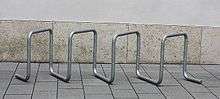 A free-standing bike rack, consisting of galvanized steel tubing bent into multiple square loops set at 45 degrees, on concrete slabs in front of a concrete wall