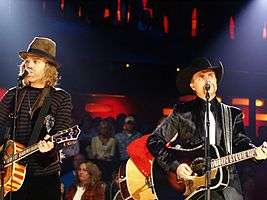 Two men playing guitars and singing into microphones, one in a brown felt hat and striped top and the other in a black cowboy hat and dark jacket