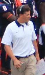 Waist-up photograph shot from a high angle of Lazor on a football sideline wearing a white polo shirt with a Nike logo, tan pants and a headset and holding a play sheet in his left hand and a marker in his right