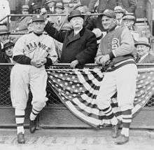 At a baseball stadium, two men in baseball uniforms stand on the field, flanking a well-dressed man who pantomimes throwing a baseball.