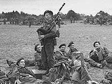 Black-and-white photograph of Millin in uniform, standing and playing his bagpipe, while uniformed soldiers lay and lounge around him on the grass in a field