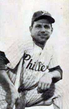 A smiling man wearing a pinstriped baseball uniform with "Phillies" across the chest and a dark baseball cap with a white "P" on the face leaning on a baseball bat
