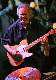 A man wearing glasses, playing a guitar and standing behind a cymbal.