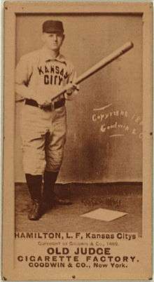 A sepia-toned image of a man in a white old-style baseball uniform and dark pillbox cap; he is holding a baseball bat in front of him in both hands
