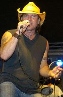 A man wearing a cowboy hat, dark T-shirt and jeans, singing into a microphone