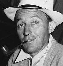 Black and white photo of Bing Crosby in 1942—a white man with light eyes wearing a light hat with a cigar in his mouth.