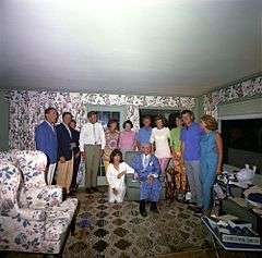 Family group portrait in sunroom with flowered drapes and upholstery with all standing behind Joe who is seated, Jackie is kneeling next to him