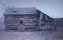 A log cabin with some logs missing and a lean-to attached to its side