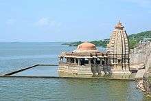 A Hindu temple with water submerged courtyard near a dam's reservoir.