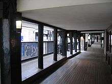 An elevated covered walkway with the fronts of premises on the right and posts and railings overlooking the street on the left