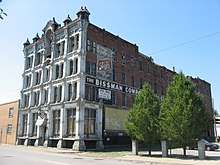 A large, red-brick building with a peeling, white-painted front. A sign on the side of the building is obscured by trees, but partially reads "The Bissman Comp..."