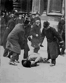 A woman is on the ground with her gloved hands over her face. A man in a top hat is holding back one police officer, while another is holding one of his gloves and stooping over the woman. In the background are several police officers and other men. Beyond them are the walls and doorway to the Parliament buildings.