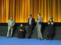 Scott Franklin, Mila Kunis, Vincent Cassel, Darren Aronofsky, and Sandra Hebron stand on a stage with a golden curtain backdrop wearing formal attire and discussing Black Swan