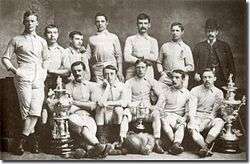 A group of twelve men, eleven in association football attire typical of the late nineteenth century and one in a suit and bowler hat.  They are displaying a number of trophies.