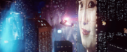 Screenshot of a police spinner flying through a cityscape next to a large building which has a huge face projected onto it. In the distance a screen can be seen with writing and pictures on it
