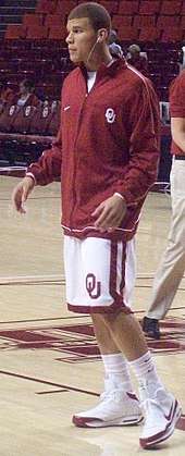 A man wearing a red jacket with white shorts with the letters "O" and "U" interlocking on the left knee and white sneakers with socks up to mid-calk. He is facing to the left of the picture.