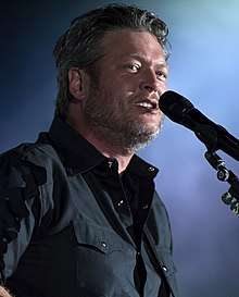 A man with greying harir and beard stubble, wearing a dark shirt, singing into a microphone
