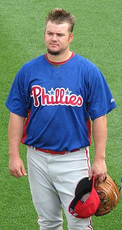 A man with spiked hair and a goatee wearing a blue baseball jersey with "Phillies" in red across the front and white baseball pants with red pinstripes stands on a baseball field holding a tan baseball glove and a red baseball cap in his left hand.