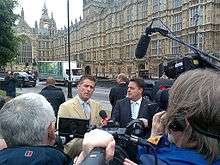 A crowd of journalists and photographers are addressed by two men, standing in front of the Palace of Westminster.