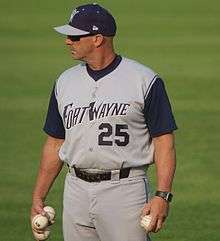 A man in a gray and navy blue baseball uniform with "Fort Wayne" on the chest stands on a baseball field holding baseballs in each hand.