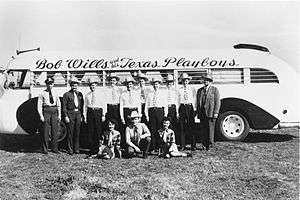 Nine men in cowboy hats, one man in a business suit, one man in a police uniform and two young women, all in front of a bus with "Bob Wills and his Texas Playboys" painted on the side