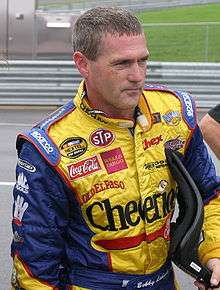 A man in his early forties wearing yellow racing overall and he is holding a HANS device