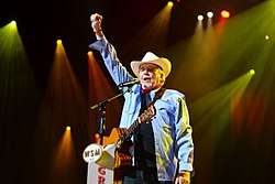 An older man wearing a white cowboy hat and a denim shirt, with a guitar hanging over his shoulder standing at a microphone raising his right fist in the air