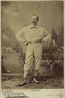 A sepia-toned photograph of a man in an old-style white baseball uniform standing with arms akimbo