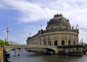 An ornate grey stone building on the point of an urbanized island. The building is connected by two bridges to the neighboring banks