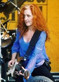 A woman with curly red hair wearing a blue shirt holding a guitar.
