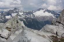 Angular light-gray rocks in the foreground. Behind, a range of darker gray mountains with snow. The one at the right has a substantial glacier at its foot.