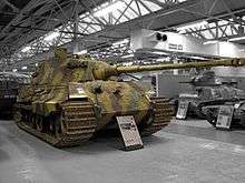 A large, turreted tank with dull yellow, green and brown wavy camouflage, on display inside Bovington museum. The tracks are wide, and the frontal armour is sloped. The long gun overhangs the bow by several meters.