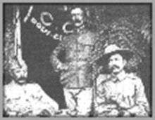 Photograph of three men at a Boy Scout event circa 1910.  The man seated on the left is unidentified, Burnham is in the middle, standing, and Baden-Powell is on the right, seated.  There is a table in front of the men.  Baden-Powell is wearing his stetson hat, Burnham has no hat, and man on the left has a modest hat.  Behind Burnham and to his right is a flag partially opened.