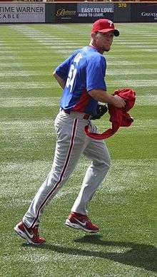 A man in a blue baseball jersey and gray baseball pants with a red stripe jogging on a baseball field