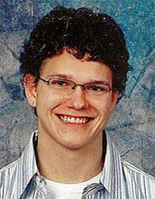 A smiling young Caucasian man with short dark brown curly hair, wearing glasses and a blue and white striped shirt with collar open, against a patterned blue background