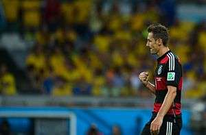 Klose pulling his arm towards himself in a goal celebration. He is wearing a red and black hooped kit. Fans in yellow Brazil jerseys are sat in the background.