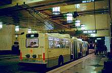 A white bus with raised poles stopped at the curb of an underground station. The granite-tiled floors reflect bright lanterns placed above the platform, adjacent to an upper level walkway.