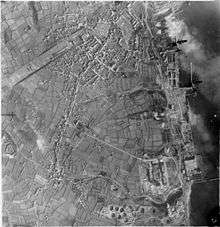 Vertical aerial photograph taken during a daylight attack on German warships docked at Brest, France. Two Handley Page Halifaxes of No. 35 Squadron RAF (upper right) fly over the naval dockyard, towards the dry docks in which the battlecruisers Scharnhorst and Gneisenau are berthed (top right) and over which a smoke screen is rapidly spreading. At middle right, a stick of bombs can be seen to have exploded inland from their intended target, Prinz Eugen, moored by the quayside. 47 aircraft from 3, 4 and 5 Groups took part in the operation, claiming accurate bombing on their targets for the loss of six aircraft