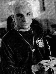 A grainy black and white photo of a bald or close-cropped man with earrings in both ears looking up slightly towards the camera, wearing a long-sleeved loose dark shirt with a crown on the left breast and a long necklace ending in a cross-like figure