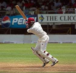 A man wearing white cricket clothes and maroon helmet plays a shot. He is standing on a cricket pitch, and the seating area of the ground is visible in the background.