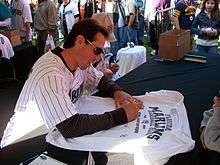 A young dark-haired man with sunglasses writing on a baseball jersey